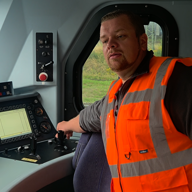 Bild des Prüfers Enrico Görzen bei der Arbeit Mann in orange Sicherheitsjacke im Führerstand eines Fahrzeugs, schaut in die Kamera.